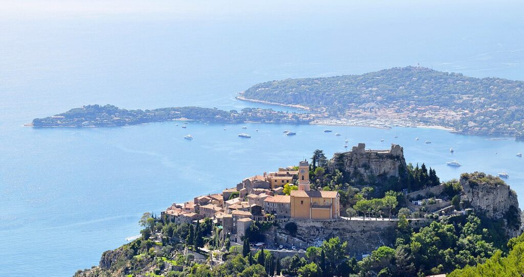 The village of Èze, with its church “Our Lady of Assumption” (Notre-Dame de l’Assomption) and its castle in the foreground, and the Cape Ferrat in the background. | Photo by Tobi 87 (CC-BY-SA-3.0)